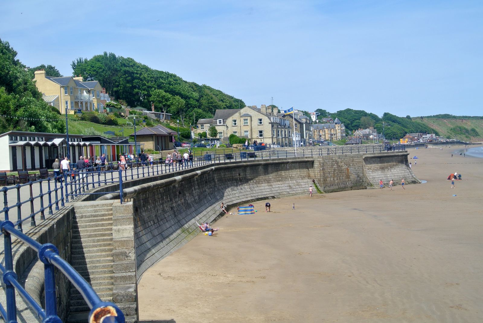 Filey seafront in summer: the curved sea wall and promenade with families on the beach and Victorian terraces on the wooded hillside